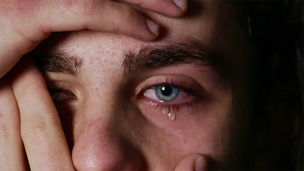 A man with tears coming out of his eye covers his face.